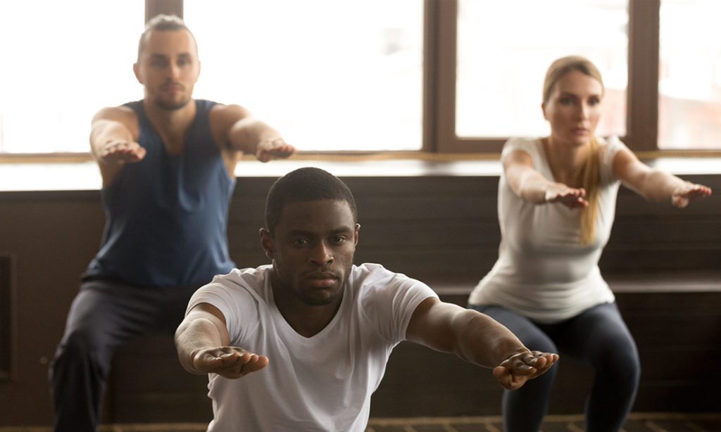 Young african american man doing squat exercise at group fitness training, sporty black guy focused on self-improvement working out with diverse active people in gym studio during routine session